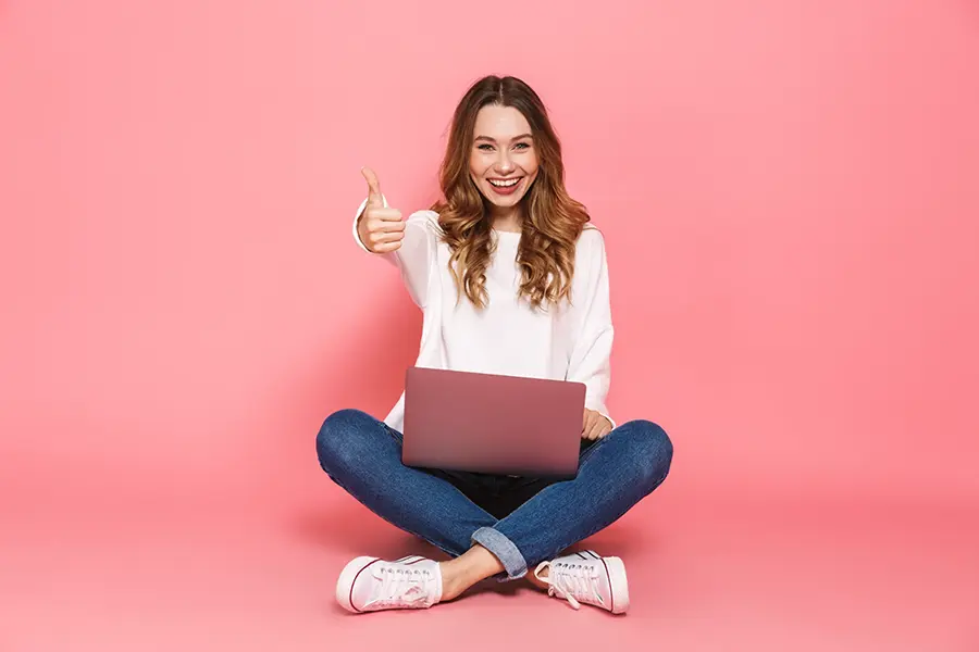 Smiling woman sitting cross-legged with a laptop on her lap, giving a thumbs-up against a pink background.