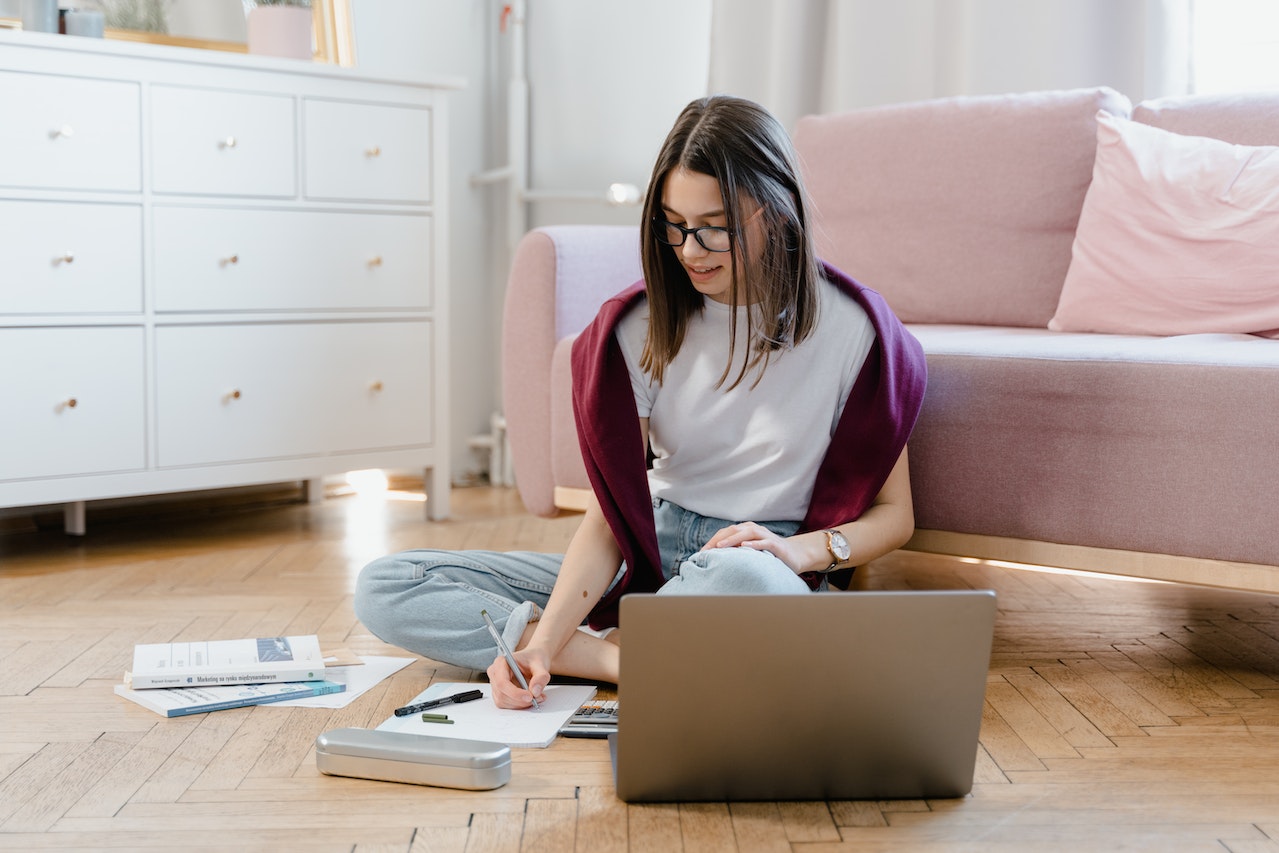 A woman sits cross-legged on the floor with a laptop open in front of her, focused on her studies. She is engaged in elearning or online learning.